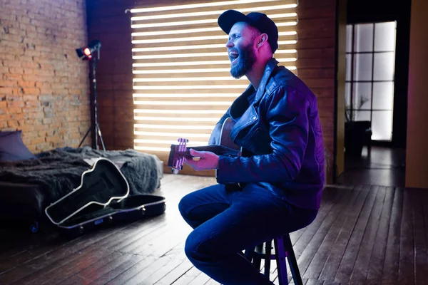 Singer and guitar player singing on a stage with neon lights. He is a rocker and he is wearing leather biker jacket or asymmetric zip jacket with black cap, jeans. — Stock Photo, Image