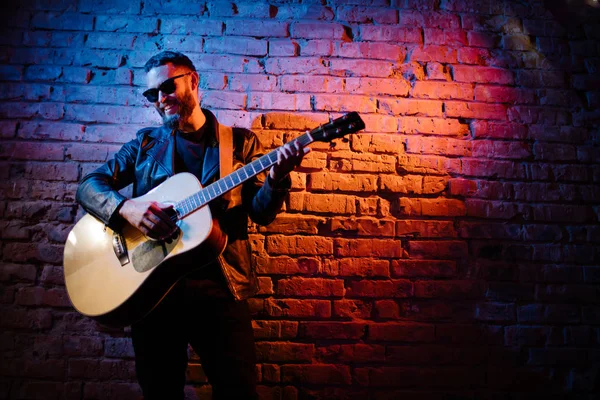 Sänger und Gitarrist singen auf einer Bühne mit Neonlichtern. er ist Rocker und trägt eine Motorradjacke aus Leder oder eine asymmetrische Reißverschlussjacke mit schwarzer Kappe, Jeans. — Stockfoto