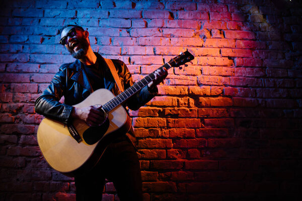 Singer and guitar player singing on a stage with neon lights. He is a rocker and he is wearing leather biker jacket or asymmetric zip jacket with black cap, jeans.