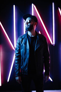 Şehir sokaklarında hipster yakışıklı adam Neon işaretler tarafından aydınlatılmış olmak. O siyah kap, kot pantolon ve güneş gözlüğü ile deri motosikletçi ceket veya asimetrik fermuar ceket giyiyor.