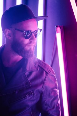 Şehir sokaklarında hipster yakışıklı adam Neon işaretler tarafından aydınlatılmış olmak. O siyah kap, kot pantolon ve güneş gözlüğü ile deri motosikletçi ceket veya asimetrik fermuar ceket giyiyor.