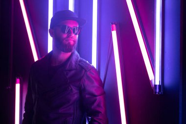 Şehir sokaklarında hipster yakışıklı adam Neon işaretler tarafından aydınlatılmış olmak. O siyah kap, kot pantolon ve güneş gözlüğü ile deri motosikletçi ceket veya asimetrik fermuar ceket giyiyor.