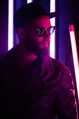 Şehir sokaklarında hipster yakışıklı adam Neon işaretler tarafından aydınlatılmış olmak. O siyah kap, kot pantolon ve güneş gözlüğü ile deri motosikletçi ceket veya asimetrik fermuar ceket giyiyor.