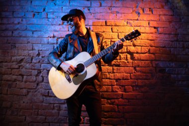 Şarkıcı ve gitarist neon ışıkları ile bir sahnede şarkı. O bir rockçı ve o deri bisikletçi ceket veya siyah kap, kot pantolon ile asimetrik zip ceket giyiyor