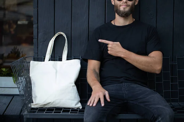 Young man holding white textile eco bag against urban city background. Ecology or environment protection concept. White eco bag for mock up. — Stock Photo, Image