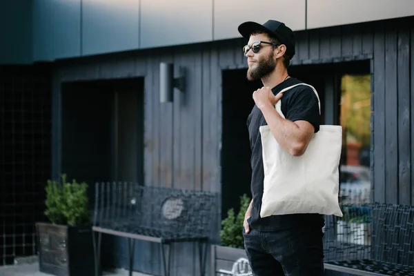 Young man holding white textile eco bag against urban city background. Ecology or environment protection concept. White eco bag for mock up. — Stock Photo, Image