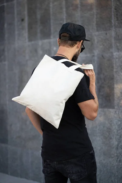 Junger Mann mit weißer Textil-Öko-Tasche vor städtischem Hintergrund. Ökologie oder Umweltschutzkonzept. weiße Öko-Tasche für Attrappen. — Stockfoto