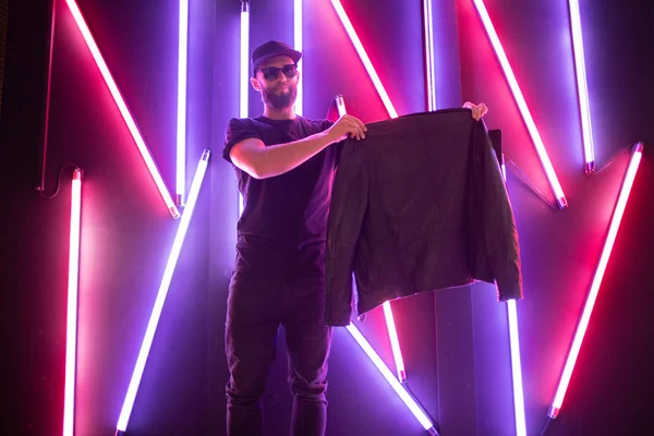 Hipster gutaussehender Mann auf den Straßen der Stadt, beleuchtet von Leuchtreklamen. er trägt Biker-Lederjacke oder asymmetrische Reißverschlussjacke mit schwarzer Mütze, Jeans und Sonnenbrille. — Stockfoto