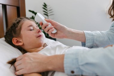 Anne, bir çocuk kızın sıcaklığını kontrol ediyor. Çocuk yatakta yatarken hasta.