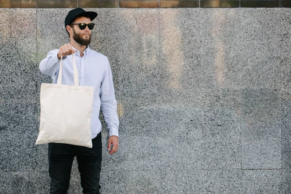 Jonge man Holding witte textiel Eco tas tegen stedelijke stad achtergrond. Ecologie of milieubescherming concept. Witte Eco-tas voor mock up. — Stockfoto