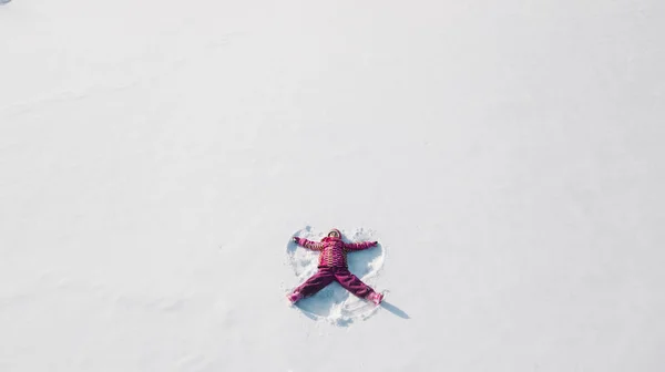 雪の天使を作る雪の上の子供。上空からの空中写真。彼女は赤い服を着ている — ストック写真