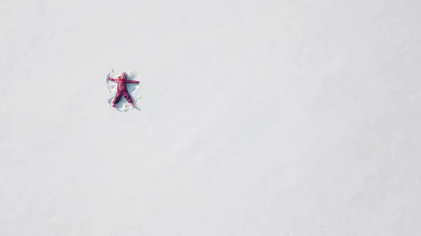 Ένα παιδί σε ένα χιόνι που φτιάχνει έναν άγγελο χιονιού. Εναέρια βολή από πάνω. Φοράει κόκκινα ρούχα. — Φωτογραφία Αρχείου