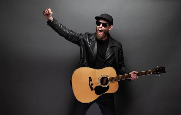 Cantante y guitarrista cantando en un escenario. Él es un rockero y lleva chaqueta de cuero motorista o chaqueta con cremallera asimétrica con gorra negra, jeans . —  Fotos de Stock