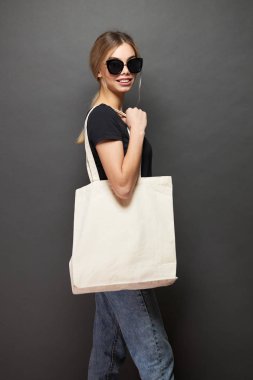 Gri arka plan karşı beyaz tekstil eko çanta tutan genç kadın. Ekoloji veya çevre koruma kavramı. Sahte için beyaz eko çanta.
