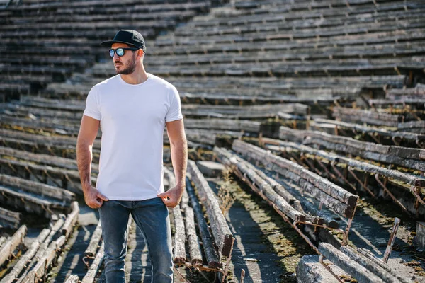 Hipster handsome male model with beard wearing white blank t-shirt and a baseball cap with space for your logo or design in casual urban style — Stock Photo, Image