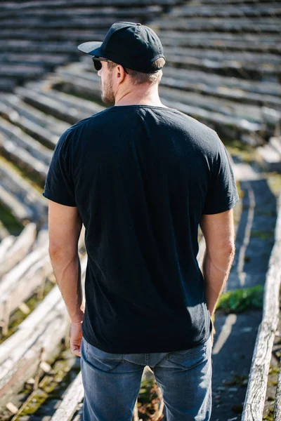 Hipster handsome male model with beard wearing black blank t-shirt and a baseball cap with space for your logo or design in casual urban style — Stock Photo, Image