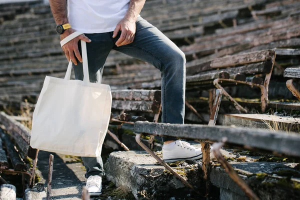 Joven sosteniendo bolso ecológico textil blanco contra fondo urbano. Ecología o concepto de protección del medio ambiente. Bolso ecológico blanco para maqueta . —  Fotos de Stock