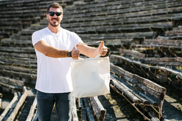 Jonge man Holding witte textiel Eco tas tegen stedelijke stad achtergrond. Ecologie of milieubescherming concept. Witte Eco-tas voor mock up. — Stockfoto