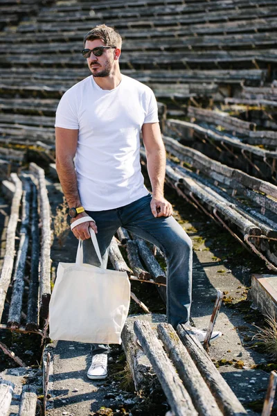 Young man holding white textile eco bag against urban city background. Ecology or environment protection concept. White eco bag for mock up. — Stock Photo, Image