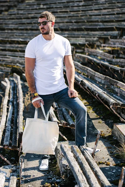 Ung man innehar vit textil Eco Bag mot Urban City bakgrund. Ekologin eller miljöskydds konceptet. Vit Eco Bag för mock-up. — Stockfoto