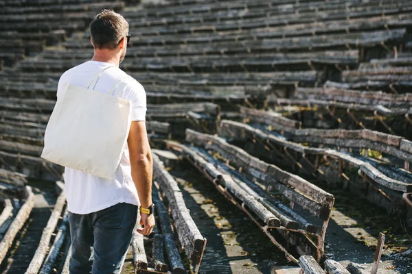 Młody mężczyzna trzyma białą tekstylne eko worek przed miejskim tle miasta. Ekologia lub koncepcja ochrony środowiska. Biała torba eko do makiety. — Zdjęcie stockowe