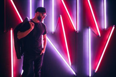 Şehir sokaklarında hipster yakışıklı adam Neon işaretler tarafından aydınlatılmış olmak. O siyah kap, kot pantolon ve güneş gözlüğü ile deri motosikletçi ceket veya asimetrik fermuar ceket giyiyor.