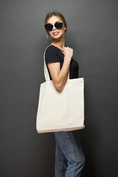 Mujer joven sosteniendo bolso ecológico textil blanco sobre fondo gris. Ecología o concepto de protección del medio ambiente. Bolso ecológico blanco para maqueta . — Foto de Stock