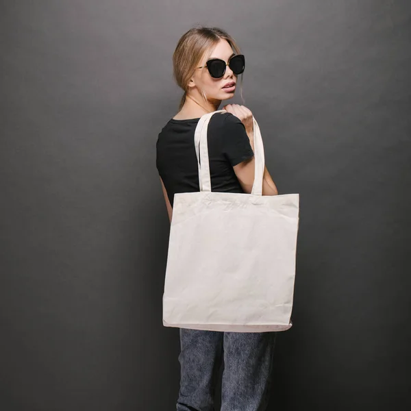 Mujer joven sosteniendo bolso ecológico textil blanco sobre fondo gris. Ecología o concepto de protección del medio ambiente. Bolso ecológico blanco para maqueta . — Foto de Stock