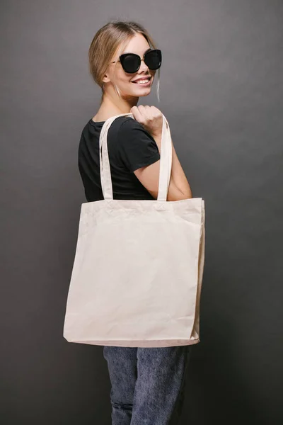 Young woman holding white textile eco bag against gray background. Ecology or environment protection concept. White eco bag for mock up. — Stock Photo, Image