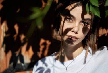 Makyajlı seksi esmer kadın dışarıda beyaz elbiseler giyiyor ve yüzünde ağaç gölgeleri var.