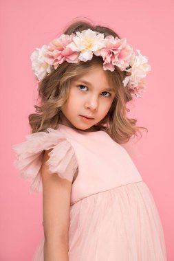 Kafasında çiçek çelengi ve bukleleri olan küçük güzel bir kızın stüdyo fotoğrafı. Arka plan, aksesuarlar ve pembe elbiseler. Kuaför salonu. Çocuk kozmetiği. Ciddi özenli çocuk.