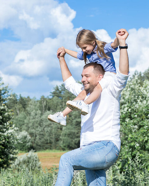 A father carries his little daughter on his shoulders ana hand against the blue sky. Outdoor walks. They are happy together, family relations, parents love for children