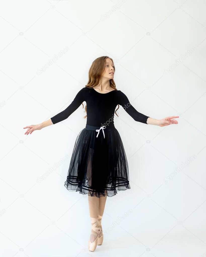 A young beautiful ballerina girl stands in motion on Pointe shoes in a black swimsuit and skirt with light background. Full-length photo. Studio of modern choreography, ballet, dance