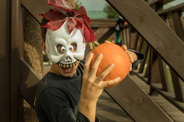 A masked boy with a pumpkin as a Ghost celebrates Halloween among wooden planks. Positive emotions. Close-up portrait, laughing face of a horror story