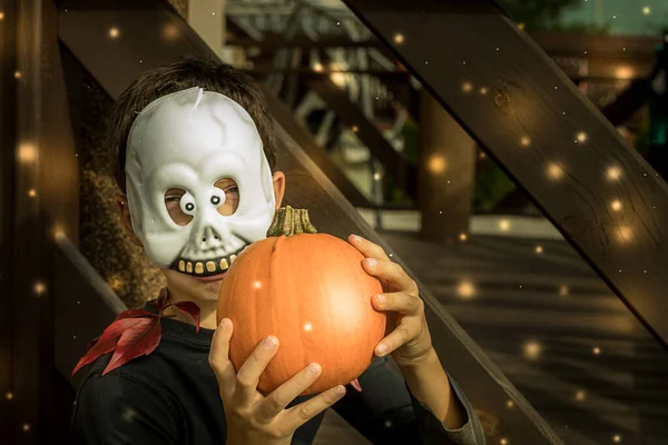 A boy in a mask with a pumpkin in the role of a Ghost celebrates the Halloween holiday among wooden boards and fairy-tale glowing lights. Positive emotions. Close-up portrait of smiling mask
