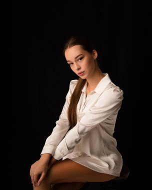 Güzel esmer kadın seksi model, vücudunda beyaz ipek gömlek var. Zarif kalça ve diz. Stüdyodaki siyah arkaplandaki çerçeveye bakıyor. Portre