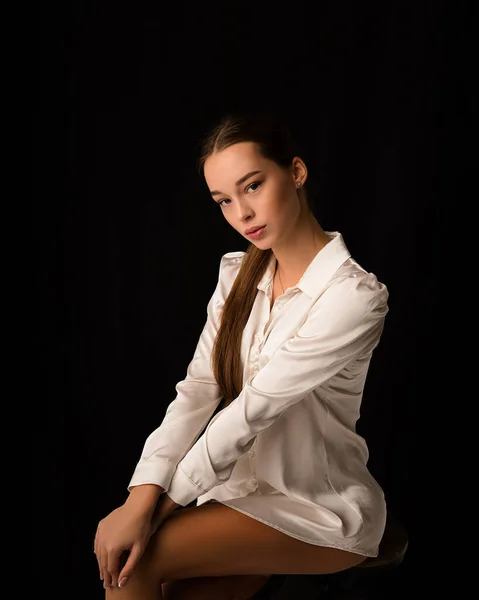 Bella ragazza bruna modello sexy, vestita con una camicia di seta bianca sul corpo. Elegante anca e ginocchio. Guarda nella cornice su uno sfondo nero nello studio. Ritratto — Foto Stock