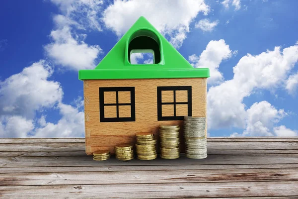 Saving money for house concept, with symbolic wooden house and pile of money against clear blue sky