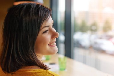 Sarı kazaklı güzel bir esmer restoranda gülümsüyor. Pencerenin kenarındaki kafede gülümseyen güzel kız.