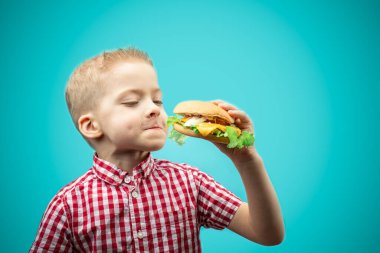 Çocuk hamburger yiyor. Çocuklar hamburgere bayılır. Mavi arka planda kırmızı gömlekli çocuk.