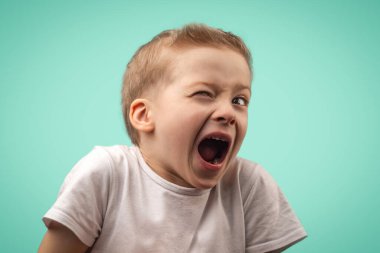 Beyaz tişörtlü çocuk bir gözünü kırpıyor. Mavi arka planda yaramaz bir çocuk