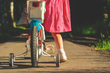 Kırmızı elbiseli küçük kız bisiklet sürmeyi öğreniyor. Öğrenmek için fazladan tekerlekleri olan çocuk bisikleti. ilk motor