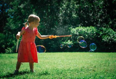 Yaz parkındaki çocuklar. Kırmızı elbiseli küçük kız parkta büyük sabun köpükleri üflüyor.