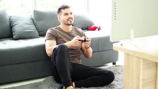 一个可爱的家伙正坐在家里的一个操场上玩电子游戏 呆在家里 男人手里拿着游戏机控制器玩 — 图库视频影像