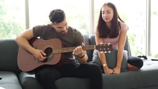 男人在客厅和妻子的女朋友一起弹奏吉他手 男人为情人唱情歌 欢欢喜喜的情侣在客厅里一起弹奏吉他 — 图库视频影像