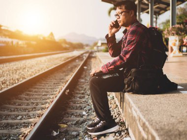 Asyalı adamın tren istasyonunda sosyal medyayı kullandığı yaz günü. Seyahat konsepti. Hipster adam telefon ve laptop kullanıyor sırt çantasıyla oturuyor ve trenin seyahat etmesini bekliyor..