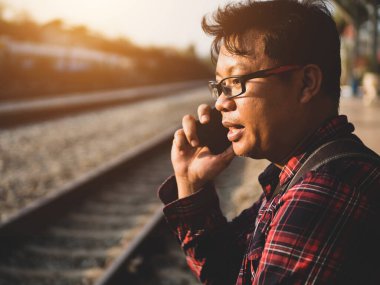 Asyalı adamın tren istasyonunda sosyal medyayı kullandığı yaz günü. Seyahat konsepti. Hipster adam telefon ve laptop kullanıyor sırt çantasıyla oturuyor ve trenin seyahat etmesini bekliyor..