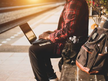 Asyalı adamın tren istasyonunda sosyal medyayı kullandığı yaz günü. Seyahat konsepti. Hipster adam telefon ve laptop kullanıyor sırt çantasıyla oturuyor ve trenin seyahat etmesini bekliyor..