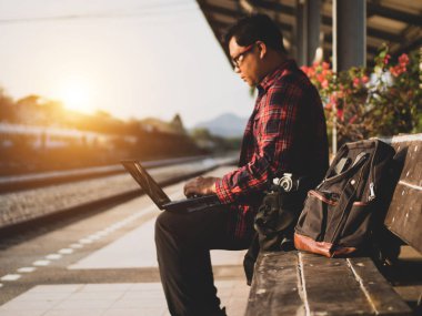 Asyalı adamın tren istasyonunda sosyal medyayı kullandığı yaz günü. Seyahat konsepti. Hipster adam telefon ve laptop kullanıyor sırt çantasıyla oturuyor ve trenin seyahat etmesini bekliyor..