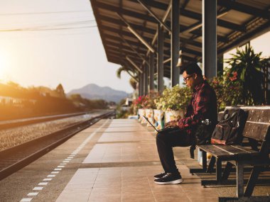 Asyalı adamın tren istasyonunda sosyal medyayı kullandığı yaz günü. Seyahat konsepti. Hipster adam telefon ve laptop kullanıyor sırt çantasıyla oturuyor ve trenin seyahat etmesini bekliyor..
